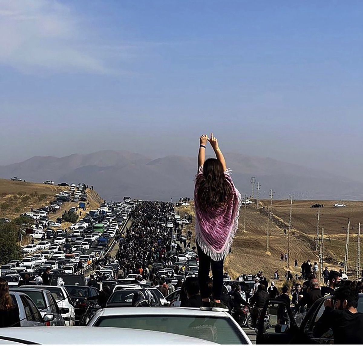 Marie Sumalla & Ghazal Golshiri, A young woman without a hijab stands on a vehicle as thousands of people make their way to the Aychi cemetery, to commemorate the 40th day of Mahsa Amini’s death, in Saqqez, her hometown in Iranian Kurdistan. Muslim tradition celebrates this date as the day of the soul’s passage to the afterlife, and the end of mourning, Saggez, Iranian Kurdistan, October 26, 2022, © Anonymous Author
